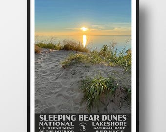 May include: A framed poster featuring a sunset over the Sleeping Bear Dunes National Lakeshore. The image shows sandy dunes with green grasses in the foreground, a calm lake, and a bright sun on the horizon. Text at the bottom reads: SLEEPING BEAR DUNES NATIONAL LAKESHORE U.S. DEPARTMENT OF THE INTERIOR NATIONAL PARK SERVICE.