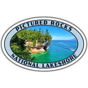 May include: Oval sticker with a blue and white border. The sticker features a scenic image of a rocky cliff with green trees and a turquoise blue lake. The text "Pictured Rocks National Lakeshore" is printed in black around the image.