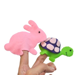 Könnte beinhalten: Fingerpuppen aus rosa Filz in Form eines Kaninchens und aus grünem Filz in Form einer Schildkröte. Das Kaninchen hat schwarze Augen und die Schildkröte hat einen lila-weißen Panzer. Die Puppen sind für das Spiel auf Fingern konzipiert.