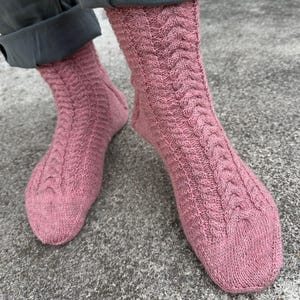 Puede incluir: Primer plano de dos calcetines rosas tejidos con un diseño de punto trenzado. Los calcetines son de un tono rosa polvoriento y parecen hechos a mano. Se llevan con pantalones grises, destacando el patrón texturizado.