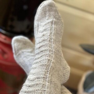 Puede incluir: Un par de calcetines de punto gris claro con un patrón texturizado. Los calcetines están doblados uno sobre otro, mostrando el detallado trabajo de puntadas. Los calcetines parecen estar hechos de un material suave y cálido.