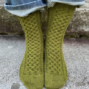 Puede incluir: Un par de calcetines de punto verde oliva con un diseño de punto de cable. Los calcetines son visibles desde los tobillos hacia abajo, con los puños de unos vaqueros azules encima. Los calcetines están hechos de un hilo suave y texturizado.