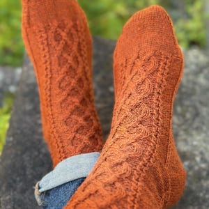Puede incluir: Un par de calcetines de punto naranja con un diseño de trenzas. Los calcetines se llevan con vaqueros azules, con los puños de los vaqueros doblados. Los calcetines son de un tono cálido y terroso.