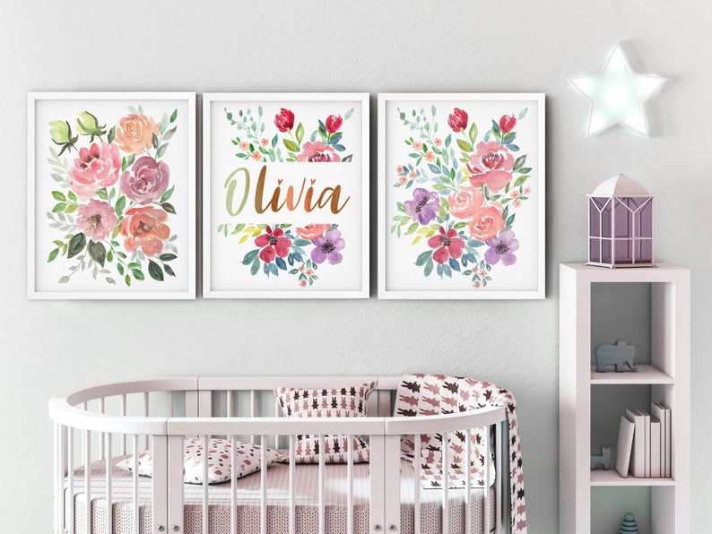 Pu&ograve; includere: Tre stampe ad acquerello incorniciate con mazzi di fiori e il nome Olivia in lettere dorate. Le stampe sono appese a un muro bianco sopra una culla bianca.