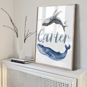 May include: A watercolor print of two whales with the name Carter written in cursive below. The whales are blue and gray and are swimming in a light blue ocean.