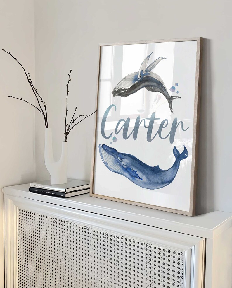 May include: A watercolor print of two whales with the name Carter written in cursive below. The whales are blue and gray and are swimming in a light blue ocean.