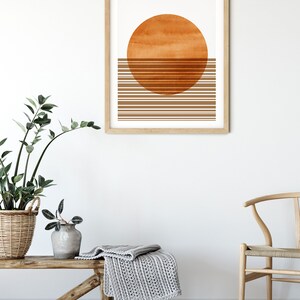 Pu&ograve; includere: Una stampa incorniciata con un design minimalista che presenta un grande cerchio arancione e linee orizzontali marroni. La stampa &egrave; appesa a una parete bianca sopra una panca di legno con una coperta di maglia grigia.