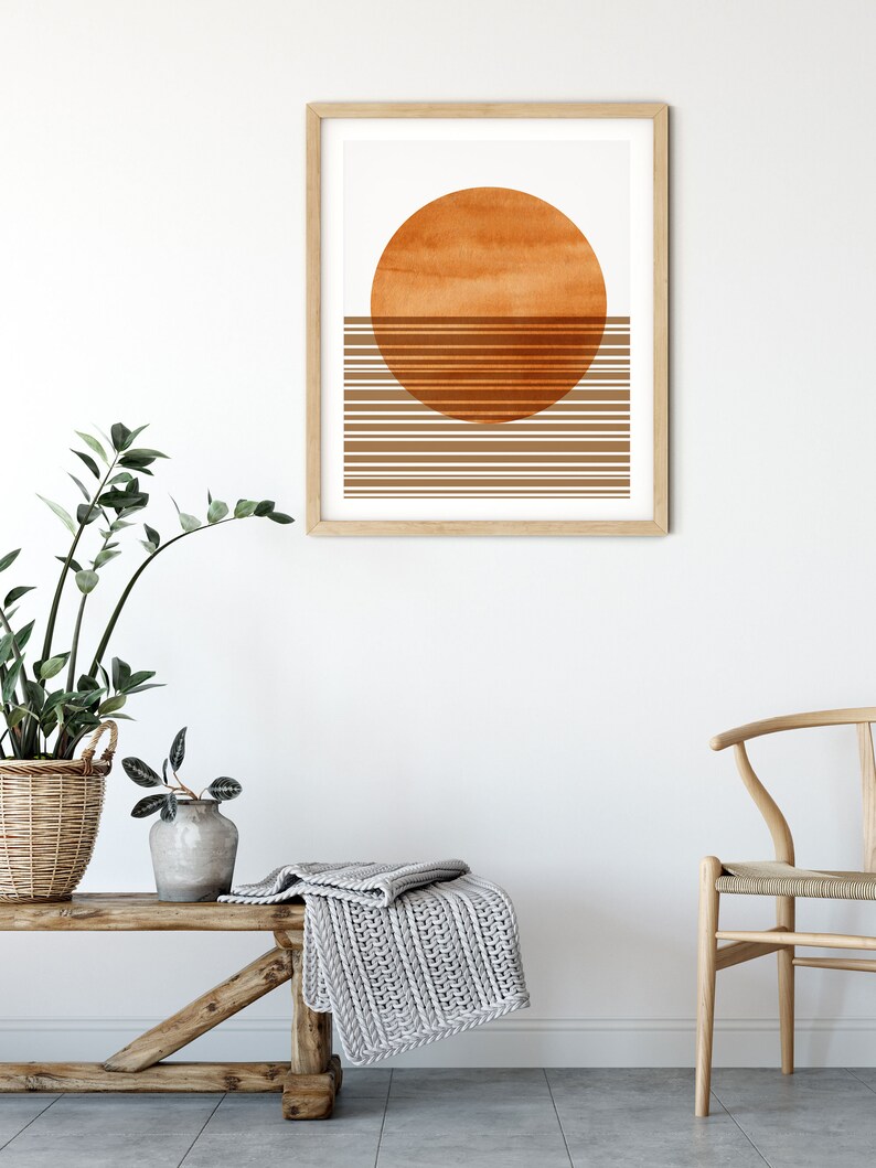 Pu&ograve; includere: Una stampa incorniciata con un design minimalista che presenta un grande cerchio arancione e linee orizzontali marroni. La stampa &egrave; appesa a una parete bianca sopra una panca di legno con una coperta di maglia grigia.