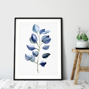 May include: A framed watercolor print featuring a sprig of blue-green eucalyptus leaves. The artwork is set against a white background, framed in black, and leans against a light gray wall. A wooden stool with a vase of greenery sits nearby.