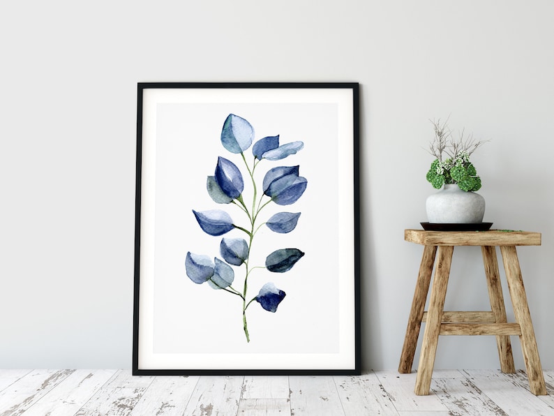 May include: A framed watercolor print featuring a sprig of blue-green eucalyptus leaves. The artwork is set against a white background, framed in black, and leans against a light gray wall. A wooden stool with a vase of greenery sits nearby.