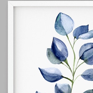 May include: A framed watercolor print featuring a sprig of blue-green eucalyptus leaves. The leaves are painted in shades of blue and green, with a soft, watercolor effect. The artwork is set against a white background and framed in a white frame.