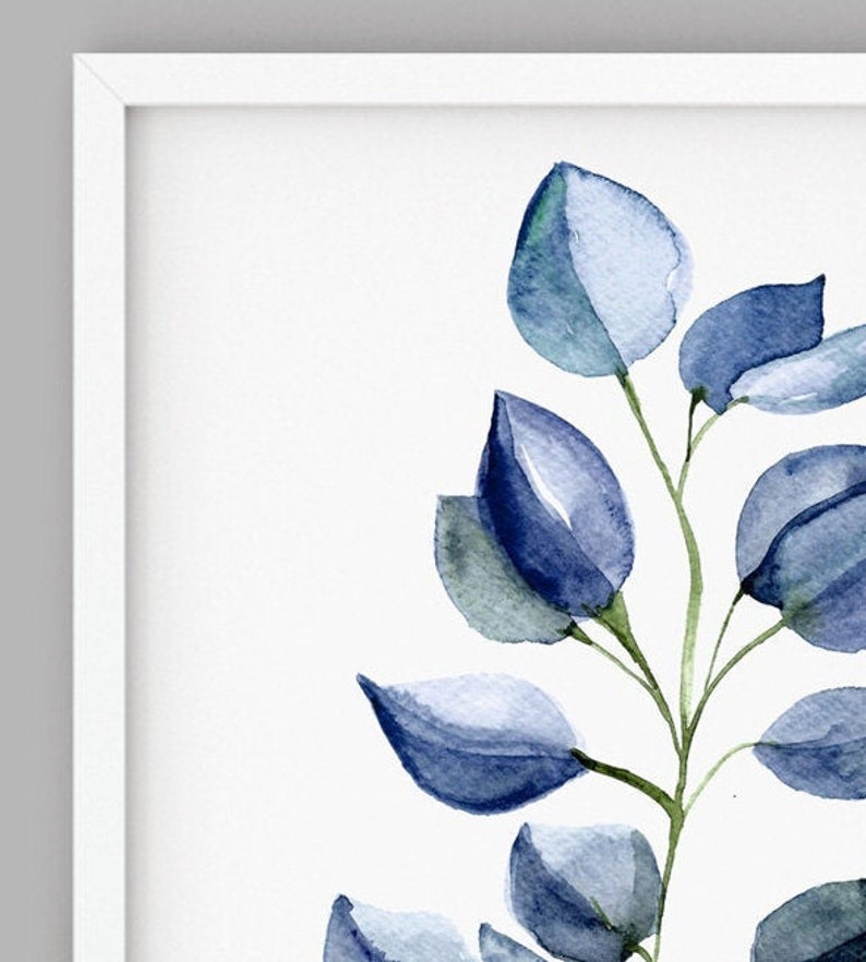 May include: A framed watercolor print featuring a sprig of blue-green eucalyptus leaves. The leaves are painted in shades of blue and green, with a soft, watercolor effect. The artwork is set against a white background and framed in a white frame.