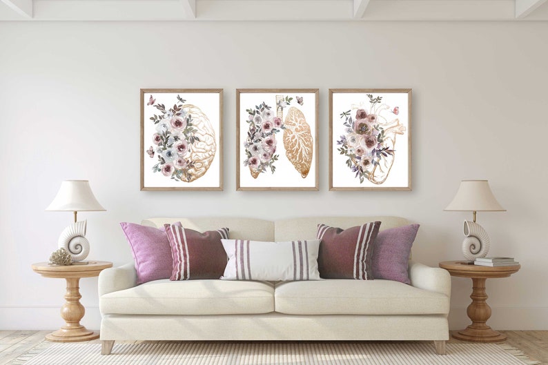 K&ouml;nnte beinhalten: Drei gerahmte Drucke mit anatomischen Illustrationen von Gehirn, Lunge und Herz, jeweils verziert mit zarten Aquarellblumen in Rosa-, Lila- und Wei&szlig;t&ouml;nen.