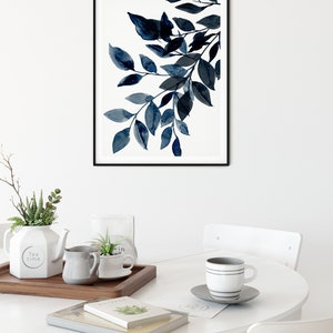 May include: A framed print of a watercolor painting of blue leaves on a white background. The print is hanging on a white wall above a white table with a cup of coffee, a teapot, and a tray with plants.