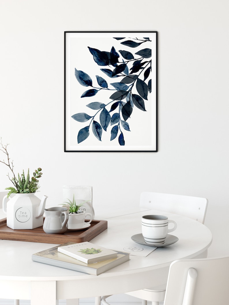 May include: A framed print of a watercolor painting of blue leaves on a white background. The print is hanging on a white wall above a white table with a cup of coffee, a teapot, and a tray with plants.