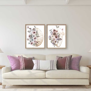 K&ouml;nnte beinhalten: Zwei gerahmte Drucke von anatomischen Illustrationen eines Gehirns und eines Herzens, jeweils mit Aquarellblumen in Rosa-, Lila- und Wei&szlig;t&ouml;nen.