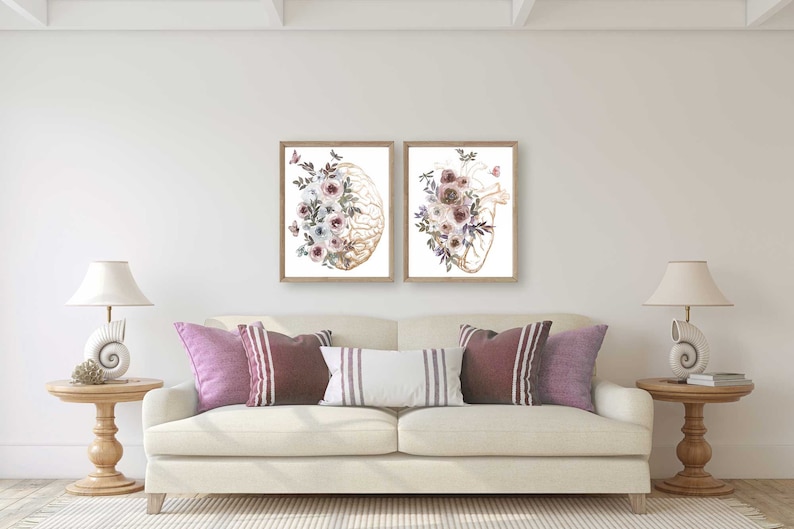 K&ouml;nnte beinhalten: Zwei gerahmte Drucke von anatomischen Illustrationen eines Gehirns und eines Herzens, jeweils mit Aquarellblumen in Rosa-, Lila- und Wei&szlig;t&ouml;nen.