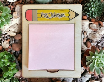 May include: A wooden desk organizer with a yellow pencil and the text "Mrs. Johnson" on it. The organizer has a pink sticky note pad.