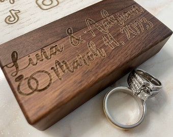 May include: A wooden ring box with the inscription "Lina & Adrian March 11, 2023". Two wedding bands are sitting on a white surface next to the box.