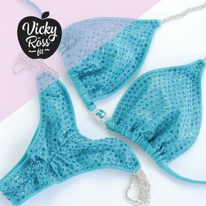 May include: Teal blue bikini with crystal embellishments and silver chains. The bikini top has a halter style and the bottom is a cheeky cut. The brand name "Vicky Ross Fit" is visible on the image.