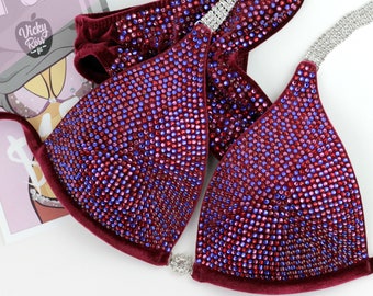 May include: Burgundy velvet bikini top and bottom with red, blue, and pink rhinestones. The bikini top has a plunging neckline and the bottom has a high-cut leg.