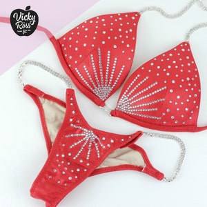 May include: A red bikini set with rhinestone embellishments. The top and bottom feature a sunburst design of rhinestones. The bikini has silver chain straps and the brand logo "Vicky Ross Fit" in the upper left corner.