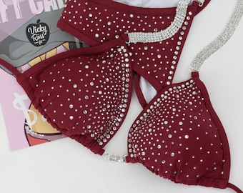 May include: Burgundy bikini with a crystal embellishment. The bikini has a halter top and a high-cut bottom. The straps are adorned with crystal chains.