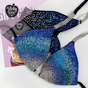 May include: A blue and black bikini top with iridescent rhinestones. The bikini top has a gradient effect, transitioning from light blue to dark blue. The straps are adorned with silver rhinestones. The image also shows a logo that says "Vicky Ross - fit".