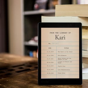 Könnte beinhalten: Ein schwarz-weißer E-Reader mit einer Bibliothekskarte auf dem Bildschirm. Die Karte ist aus der Bibliothek von Kari und listet die Buchtitel "The Fault in Our Stars", "The Alchemist", "Harry Potter and the Order of the Time", "A Court of Thorns and Roses", "Fourth Wing", "Love Theoretically" und "Funny Story" auf.
