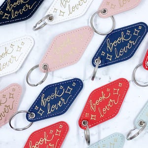 May include: A selection of colourful keyrings in shades like navy, red, and pink. Each keyring features the words "book lover" in gold script, with star accents. They are attached to silver key rings.