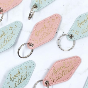 May include: A selection of pastel pink and blue keyrings, each featuring the words "book addict" in gold script. The keyrings are diamond-shaped with silver rings. The background has a white marble effect.