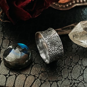 May include: A silver ring with a textured, organic design. The ring is set against a dark background with a textured surface.