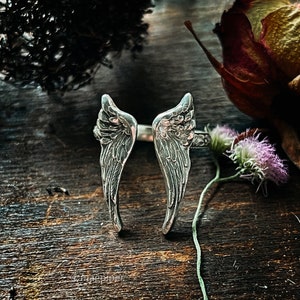 May include: A silver ring with two angel wings, the wings are detailed with textured lines. The ring is photographed on a wooden surface with dried flowers.