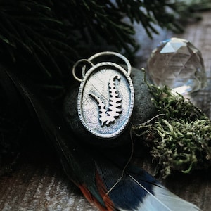 May include: A silver pendant with a fern design, set against a background of moss, a crystal, and a feather.