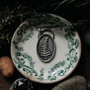 May include: A silver pendant with a fern design on a white and green patterned plate. The pendant is on a wooden surface with moss and stones.
