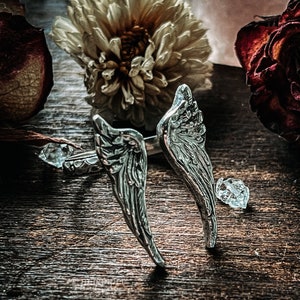 May include: Two silver angel wing rings with detailed etching on a wooden surface with dried flowers.