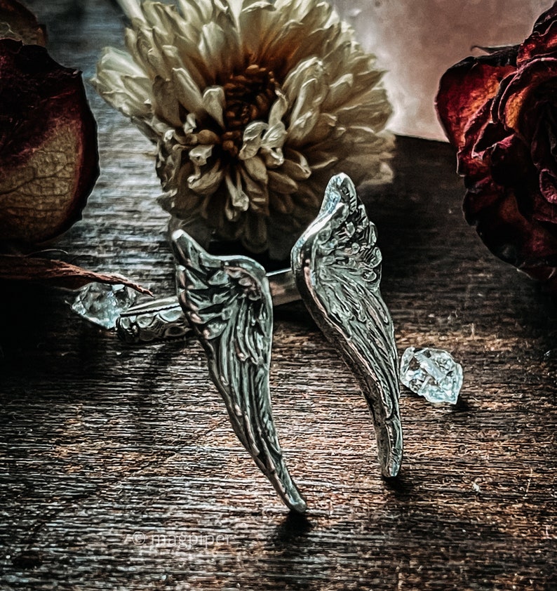 May include: Two silver angel wing rings with detailed etching on a wooden surface with dried flowers.