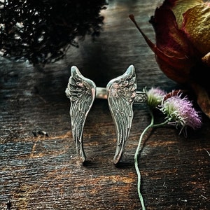 May include: A silver ring with two angel wings, the wings are detailed with a textured design. The ring is on a wooden surface with dried flowers.