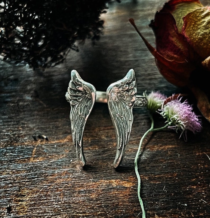 May include: A silver ring with two angel wings, the wings are detailed with a textured design. The ring is on a wooden surface with dried flowers.