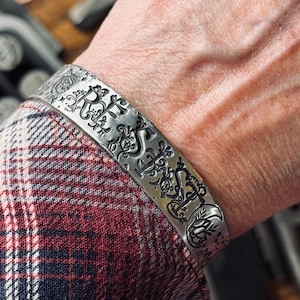 May include: A silver cuff bracelet with an ornate, engraved design. The bracelet features intricate floral and scrollwork patterns. The cuff is shown on a person's wrist, with a plaid shirt in the background.