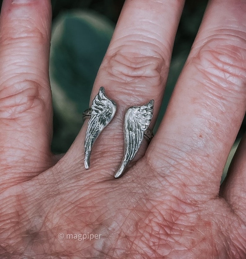 May include: A silver ring with two wings on the band. The wings are detailed and textured.
