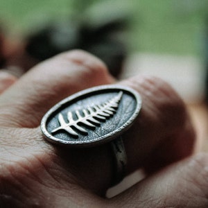 May include: A silver ring with a fern leaf design. The ring is worn on a person's finger.