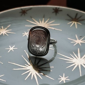 Peut inclure: Une bague en argent avec un motif orné sur le dessus. La bague est posée sur une assiette bleue et dorée avec un motif en forme d'étoile.