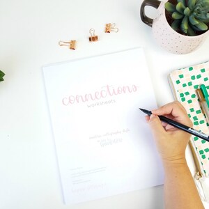May include: A white booklet with the title "Connections Worksheets" written in pink calligraphy. The booklet is open to a blank page, and a person is writing on it with a black calligraphy pen.
