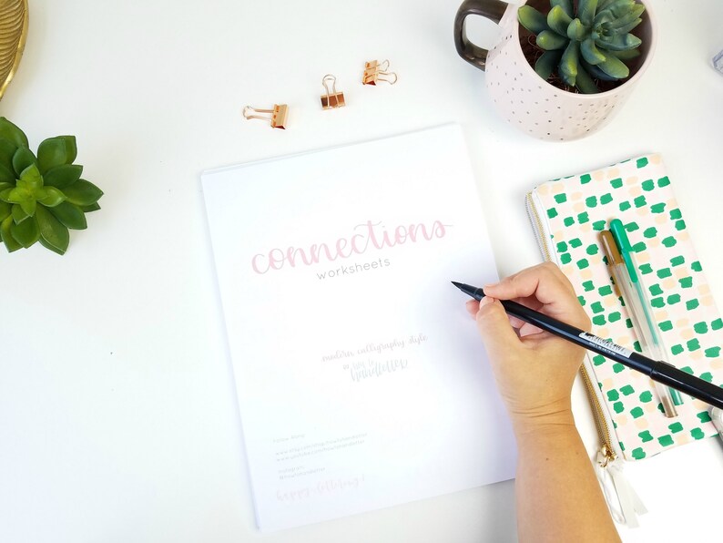 May include: A white booklet with the title "Connections Worksheets" written in pink calligraphy. The booklet is open to a blank page, and a person is writing on it with a black calligraphy pen.