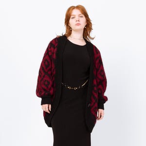 May include: A woman wearing a black dress and a black and red patterned cardigan. The cardigan has a geometric design in red on a black background. The dress is a solid black color and reaches the ankles. The woman is wearing black boots.