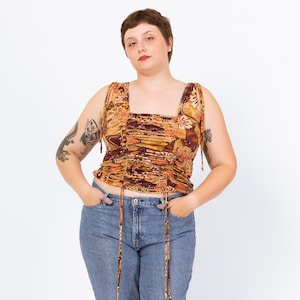 May include: A sleeveless top with a square neckline and a ruched bodice. The top features a floral pattern in shades of brown, orange, and red. The straps are thin and tie at the shoulders. The model is wearing blue jeans.