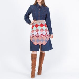 May include: A denim dress with long sleeves and a collared neckline. The dress features a colorful patchwork apron with red, green, and floral patterned squares. The outfit is completed with brown knee-high boots.