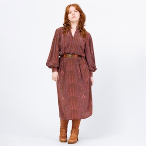 May include: A long-sleeved, burgundy dress with a paisley pattern. The dress features a V-neck, a brown belt with a decorative buckle, and long sleeves that gather at the wrists. The dress is paired with brown boots.