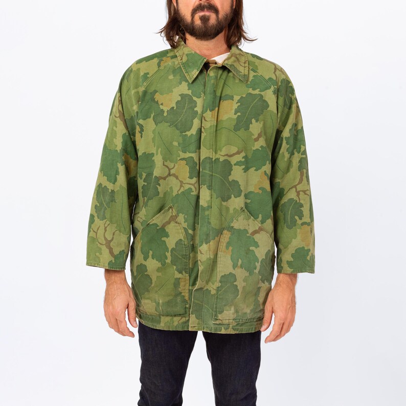 May include: Green camouflage jacket with a leaf pattern. The jacket has a collared neckline, long sleeves, and two front pockets. The jacket is made of a durable fabric and is suitable for outdoor wear. The jacket is a vintage style.